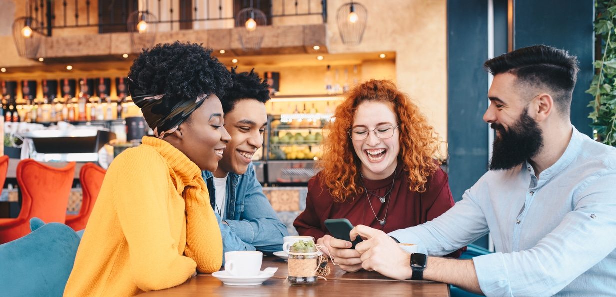 Four friends, two males and two females, sitting at a cafe, drinking coffee and talking. The bearded man is showing something on his cellphone. Building a social connection