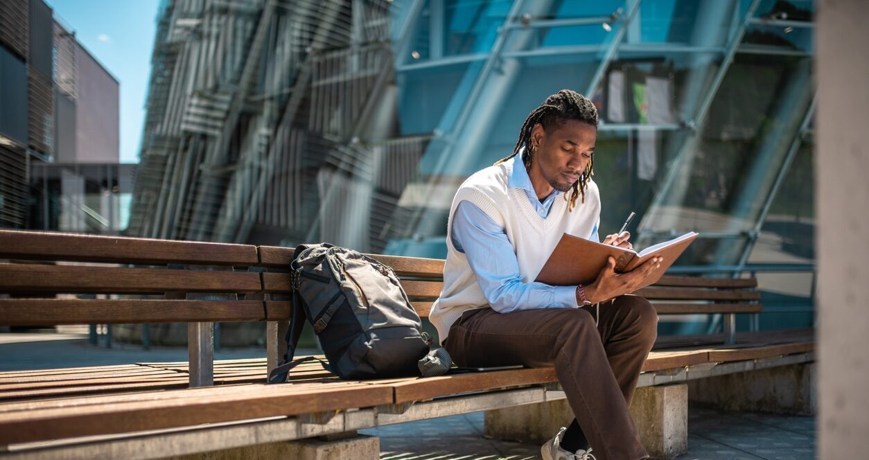 Young man sitting on a bench outside planning and setting his goals in a journal