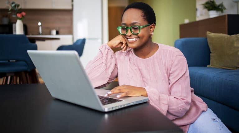 An African women sitting on the floor, however is scrolling on her laptop and is wearing some striking green glasses