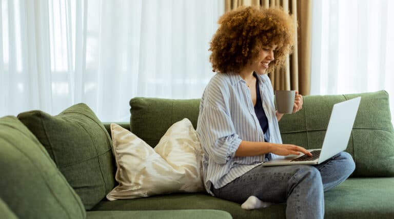 A young and youthful women sitting on her couch whilst scrolling on her laptop holding her coffee mug.